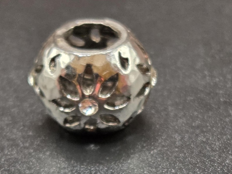 May include: A silver-colored bead with a floral design and a faceted surface. The bead has a central hole and is adorned with small, clear crystals. The bead is likely used for jewelry making or crafting.