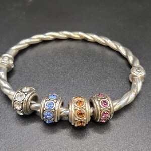 Puede incluir: Pulsera plateada con dise&ntilde;o retorcido. La pulsera presenta cuatro cuentas decorativas con piedras preciosas de colores en transparente, azul, naranja y rosa. Es una joya.