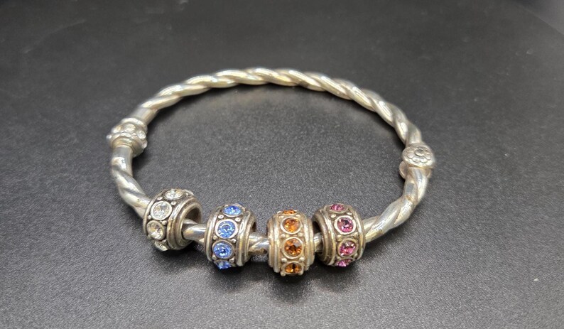 Puede incluir: Pulsera plateada con dise&ntilde;o retorcido. La pulsera presenta cuatro cuentas decorativas con piedras preciosas de colores en transparente, azul, naranja y rosa. Es una joya.