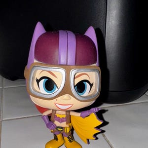 May include: A Batgirl figurine with a purple cowl and mask, brown goggles, and a yellow cape. The figure has a smiling face with blue eyes, a yellow utility belt, and yellow boots. The figure is standing on a white tiled surface.