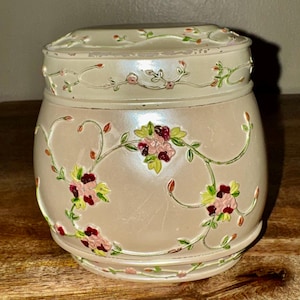 Puede incluir: Un tarro de cerámica beige con tapa, adornado con diseños florales. El tarro tiene forma redondeada y está adornado con diseños florales rosas, verdes y rojos. El diseño floral envuelve el tarro y la tapa.