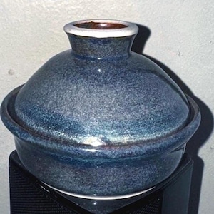 May include: A blue ceramic lidded pot with a speckled finish. The pot has a rounded body and a flared rim, with a contrasting white and brown rim on the lid. The pot is sitting on a black surface.