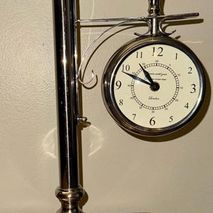 May include: A silver-colored, double-sided clock with a stand. The clock face is white with black numerals and hands. The clock reads "London" and "dream until your come true". The stand has a round base and a vertical pole.