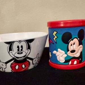 May include: A white bowl and a red-handled mug featuring Mickey Mouse. The bowl has a black and red Mickey Mouse graphic. The mug has a blue background with Mickey Mouse, yellow lightning bolts, and red accents. A fun set for kids.