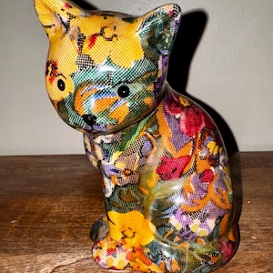 May include: A ceramic cat figurine with a colorful floral pattern. The cat is sitting with its head turned, showcasing a mix of yellow, red, purple, and green hues. The figurine has black eyes and a black nose, and is placed on a wooden surface.