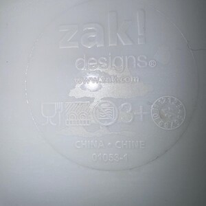 以下が含まれることがあります： 「zak! designs」と「www.zak.com」のエンボス加工が施された白いプラスチック製品。シンボルと「CHINA &bull; CHINE」と「01053-1」のテキストが含まれています。