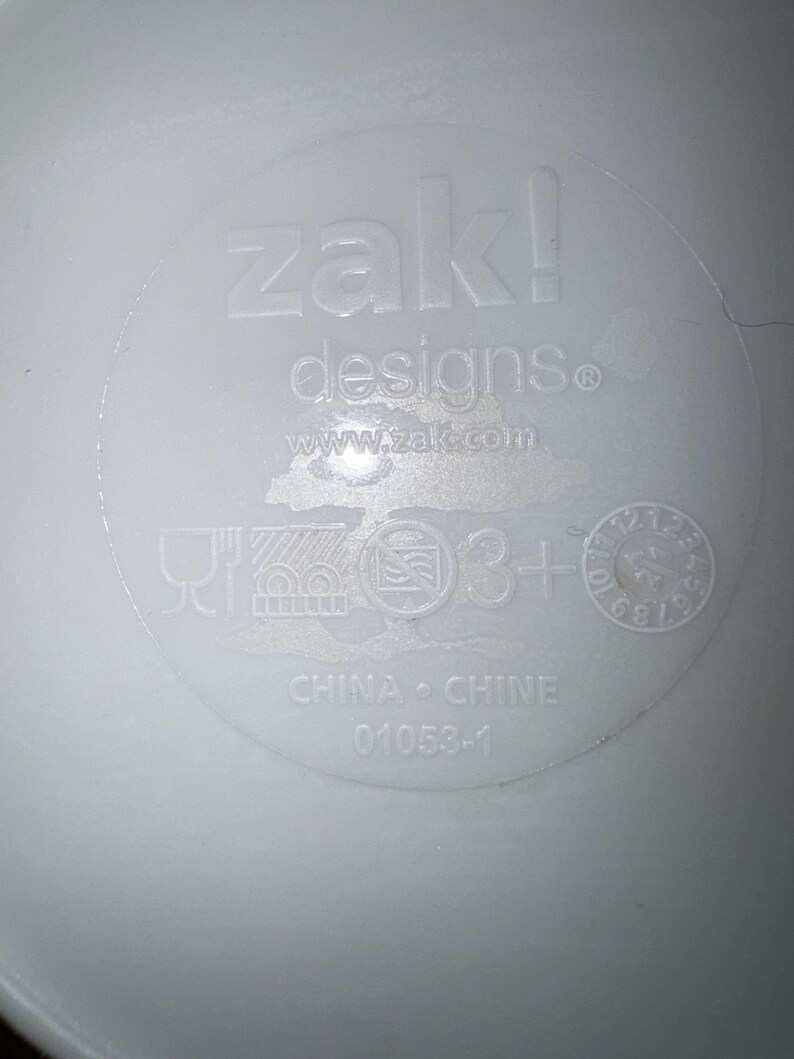以下が含まれることがあります： 「zak! designs」と「www.zak.com」のエンボス加工が施された白いプラスチック製品。シンボルと「CHINA &bull; CHINE」と「01053-1」のテキストが含まれています。