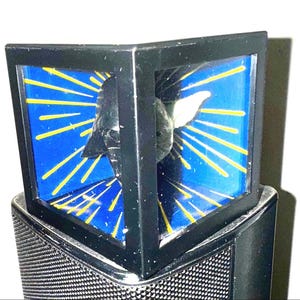 May include: A black, geometric-shaped object with a blue background and yellow rays. The object has a metallic, mesh-like base. The design features a white shape in the center, possibly a bird or a wing.