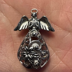May include: Silver-toned angel pendant with detailed wings and clasped hands. The pendant features a teardrop-shaped filigree design with a poinsettia flower and a white background. A small loop is at the top for hanging.