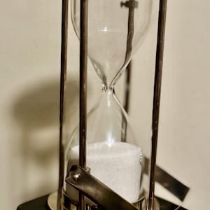 May include: An hourglass with a clear glass chamber filled with white sand. The glass is held within a metal frame with a black base. The hourglass is a decorative item, often used as a timer.