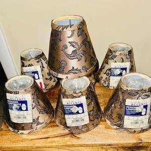 May include: Six designer lampshades with a brown and black floral pattern. The lampshades are in various sizes and shapes, with a label on each one. The lampshades are sitting on a wooden surface.