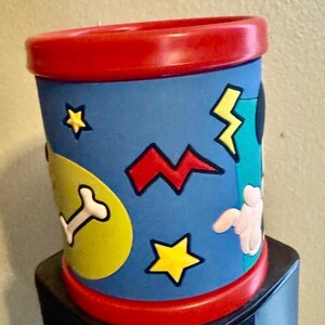 以下が含まれることがあります： 赤い縁と底を持つ円筒形の容器。青色の本体には、黄色の星、稲妻、骨などの漫画風のグラフィックが描かれています。白い手袋をした手が見えます。