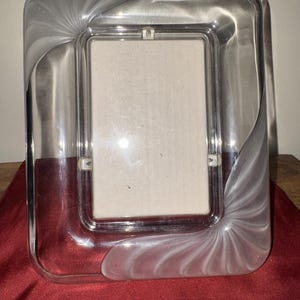 May include: A rectangular glass picture frame featuring a clear center and frosted glass corners with a swirling, fan-like pattern. The frame has a smooth, polished finish and a central rectangular opening for a photograph, with visible metal clips.