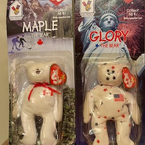 May include: Two Ty Beanie Baby bears in their original packaging. One is named Maple, with a Canadian flag on its chest and a red maple leaf. The other is Glory, with a US flag and stars. Both have the Ty heart tag.