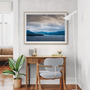 Puede incluir: Una fotografía enmarcada de un paisaje de agua y montañas bajo un cielo nublado cuelga sobre un escritorio de madera. Una silla gris con patas de madera está frente al escritorio. Una planta en maceta y una lámpara de pie blanca también están en la habitación.