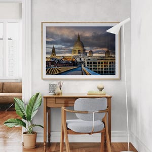 Può includere: Una stampa incorniciata della Cattedrale di St. Paul a Londra è appesa sopra una scrivania in legno. L'immagine presenta la cupola della cattedrale sotto un cielo drammatico. Una sedia moderna e una pianta in vaso sono in primo piano, creando una scena d'interni elegante.