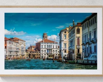 May include: A framed landscape photograph of a Venetian canal scene featuring historic multi-story buildings with light-colored facades, a central bell tower, and boats floating on the water under a bright blue sky with white clouds.