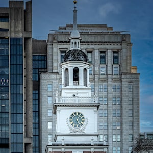 May include: A detailed photograph of a historic clock tower with a white facade and a clock face with gold hands and numerals. The tower is topped with a spire and weather vane. The background features modern and older buildings.