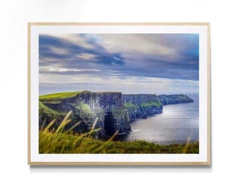 May include: A framed landscape photograph of the Cliffs of Moher, featuring steep, dark rock faces covered in green grass, overlooking a deep blue ocean under a cloudy, dramatic sky. The print is encased in a light-colored wooden frame.