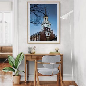 May include: A framed print of a historic building with a clock tower. The building is made of red brick and has a white clock face. The print is displayed above a wooden desk with a gray chair and a potted plant.
