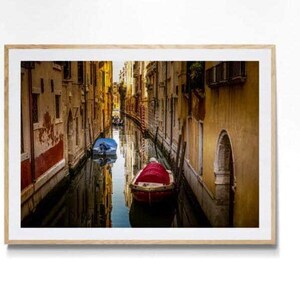 May include: A framed photograph of a Venetian canal scene. The image features a narrow waterway flanked by aged buildings with warm, golden hues. Two boats, one red and one blue, are moored in the water, reflecting the architecture.
