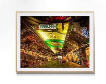 Puede incluir: Una impresión de arte mural enmarcada que representa el interior de un túnel cubierto de grafitis vibrantes. El techo presenta una iluminación verde y amarilla brillante, con el texto "JESUS IS THE" visible en pintura azul y negra.