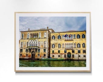 May include: A framed wall art print featuring two yellow multi-story buildings along a canal in Venice. The architecture displays arched windows, stone balconies, and weathered facades. A green boat is moored at the water level under a cloudy sky.