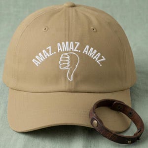 May include: A tan baseball cap featuring white embroidered text that reads "AMAZ. AMAZ. AMAZ." with a white embroidered thumbs-down icon centered below the text. A dark brown leather bracelet with metal studs rests on the brim of the cap.