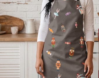 May include: A gray apron featuring a repeating pattern of nutcracker figures, ballet dancers in light blue dresses, and various colorful candies. The apron has black neck and waist straps and is worn over a long-sleeved white shirt.