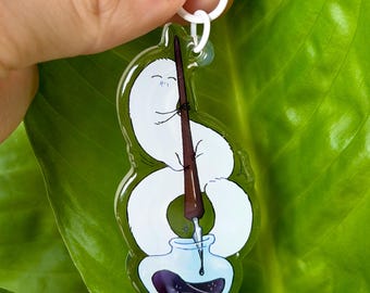 May include: A clear acrylic keychain featuring a white, worm-like creature holding a brown quill pen. The creature is positioned above a small, light blue inkwell containing dark purple ink. The keychain includes a white plastic lobster clasp.