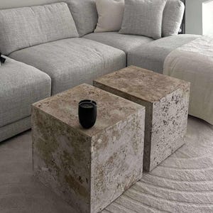 Könnte beinhalten: Zwei quadratische Beistelltische in Steinoptik mit strukturierter Oberfläche. Auf einem Tisch steht eine kleine schwarze Kerze. Die Tische stehen vor einem hellgrauen Ecksofa und einem gemusterten Teppich.
