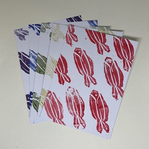 Op de afbeelding: Een stapel witte kaarten met een herhalend patroon van rode, gestileerde vogelachtige vormen. Extra kaarten tonen vergelijkbare ontwerpen in blauw, groen en paars. De kaarten zijn uitgespreid en tonen de verscheidenheid aan kleuren en patronen.