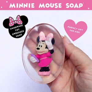 May include: Oval-shaped soap with a Minnie Mouse toy embedded inside. The soap features a pink Minnie Mouse figure wearing a pink dress and bow. The soap is held in a hand, with text that reads "MINNIE MOUSE SOAP".