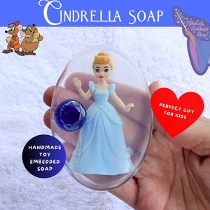 May include: Cinderella Soap with a Cinderella figurine embedded in clear soap. The soap has a blue gem and is labeled "Handmade Toy Embedded Soap". The image also includes the text "Cinderella Soap" and a red heart that says "Perfect Gift for Kids".