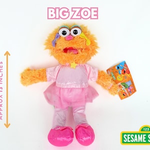May include: A plush Big Zoe doll from Sesame Street, approximately 13 inches tall. The doll is orange with large eyes, wearing a pink dress and shoes. It holds a colorful card. The Sesame Street logo is in the bottom right.