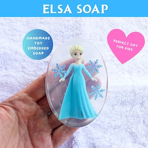 May include: Oval-shaped soap with a toy Elsa figure in a blue dress and snowflake accents. The soap is clear, with the text "ELSA SOAP" at the top. A pink heart-shaped graphic says "PERFECT GIFT FOR KIDS".