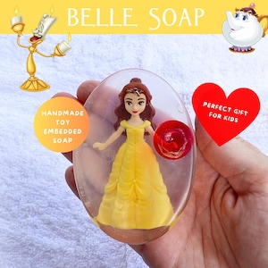 May include: A clear, oval-shaped soap bar with a Belle doll embedded inside. The doll wears a yellow gown. The soap is held in a hand. The text "BELLE SOAP" is at the top. A red heart with the text "PERFECT GIFT FOR KIDS" is also visible.