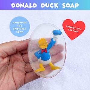 May include: Oval-shaped, clear soap with a Donald Duck toy embedded inside. The soap has the text "DONALD DUCK SOAP" at the top. A blue circle reads "HANDMADE TOY EMBEDDED SOAP". A red heart reads "PERFECT GIFT FOR KIDS".
