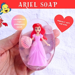 May include: Handmade Ariel soap with a toy embedded inside. The soap is clear with a pink princess figure and a seashell. The soap has the text "Ariel Soap" at the top. A red heart with the text "Perfect Gift for Kids" is also visible.