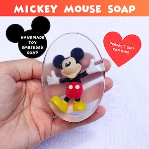 May include: Oval-shaped soap with a Mickey Mouse toy embedded inside. The soap is clear with small sparkles. The Mickey Mouse figure is wearing red shorts, yellow shoes, and white gloves. The text "Mickey Mouse Soap" is at the top.