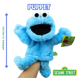 May include: A blue Cookie Monster puppet from Sesame Street, approximately 13 inches tall. The puppet has large white eyes with black pupils, an open mouth, and a plush texture. The word "PUPPET" is above the puppet, and the Sesame Street logo is visible.
