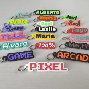 May include: A collection of colorful keychains with names and words like "GAME" and "ARCADE" in various fonts and colors. Each keychain has a silver ring for attachment.