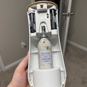 May include: An open, white automatic air freshener with two AA batteries and a Sun-Drenched Linen fragrance bottle. The bottle has a marble design and a label that reads "WHITE BARN" and "SUN-DRENCHED LINEN".