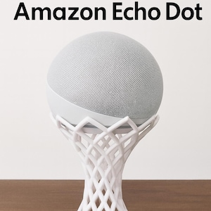May include: A white Amazon Echo Dot smart speaker on a decorative white stand. The speaker is a light gray color and has the words "Amazon Echo Dot" in black text above it. The stand has a unique, open, woven design.