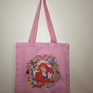 以下が含まれることがあります： ピンク色のトートバッグ。赤い髪の女性が青いトップスを着て、カラフルな花とリボンで囲まれたイラストがフレームに入っています。バッグにはお揃いのピンク色のハンドルがあり、白い壁に掛けられています。