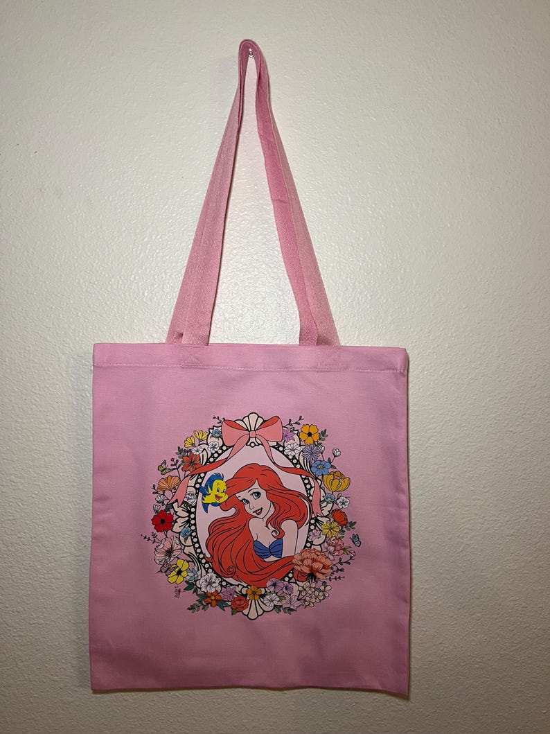 以下が含まれることがあります： ピンク色のトートバッグ。赤い髪の女性が青いトップスを着て、カラフルな花とリボンで囲まれたイラストがフレームに入っています。バッグにはお揃いのピンク色のハンドルがあり、白い壁に掛けられています。