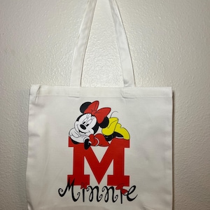May include: White canvas tote bag featuring a red letter "M" with a Minnie Mouse graphic. The design includes Minnie's face, a red bow, and the name "Minnie" in black script. The bag has white handles.