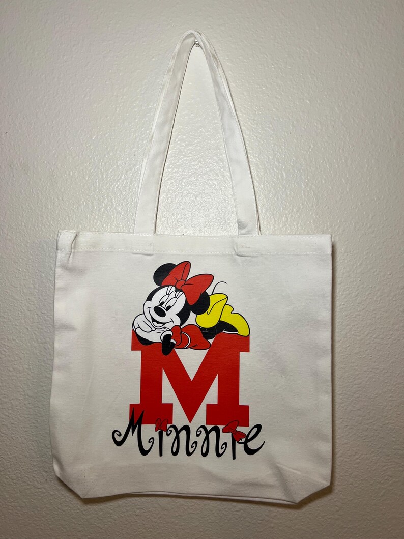 May include: White canvas tote bag featuring a red letter "M" with a Minnie Mouse graphic. The design includes Minnie's face, a red bow, and the name "Minnie" in black script. The bag has white handles.