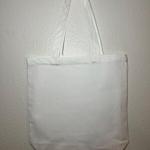May include: A plain white tote bag with two long handles. The bag is made of a sturdy fabric and hangs against a white wall. The handles are attached at the top and are the same color as the bag.
