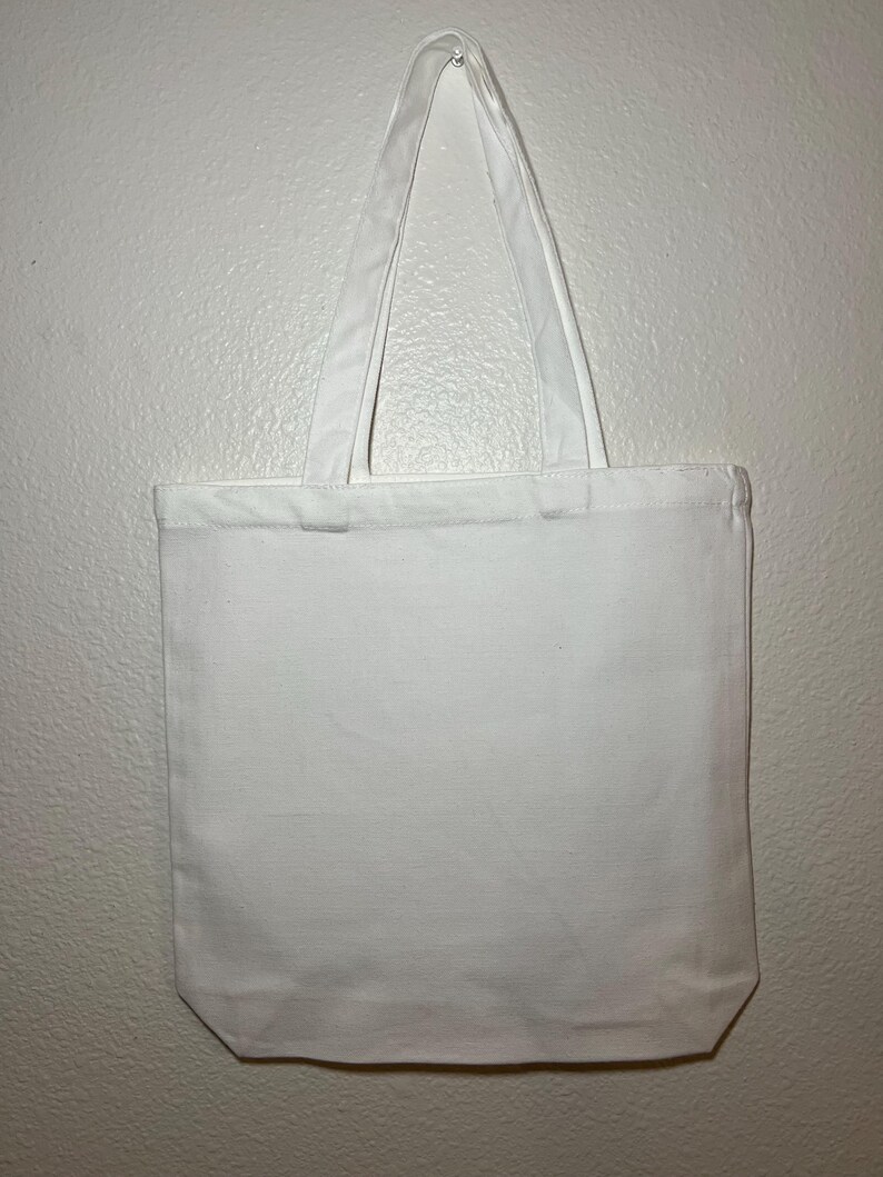 May include: A plain white tote bag with two long handles. The bag is made of a sturdy fabric and hangs against a white wall. The handles are attached at the top and are the same color as the bag.